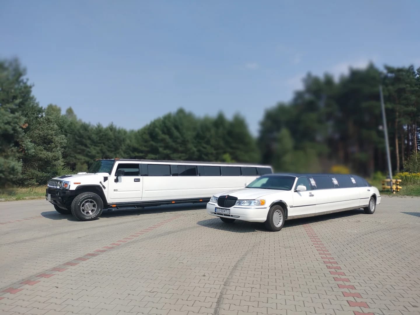 Dwie białe limuzyny, Hummer oraz Lincoln, zaparkowane obok siebie na wybrukowanym placu.