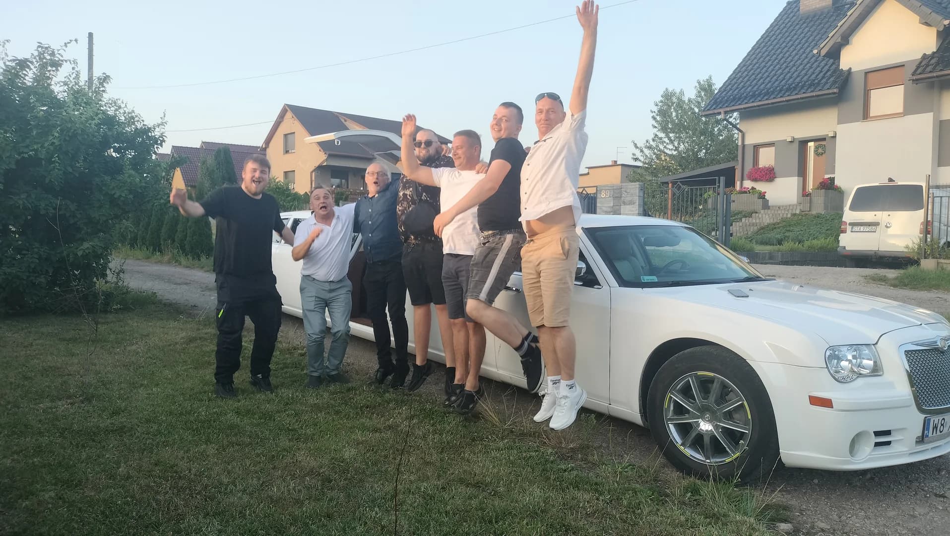 Radosna grupa mężczyzn podskakuje i świętuje obok białej limuzyny na podmiejskiej osiedlowej ulicy.