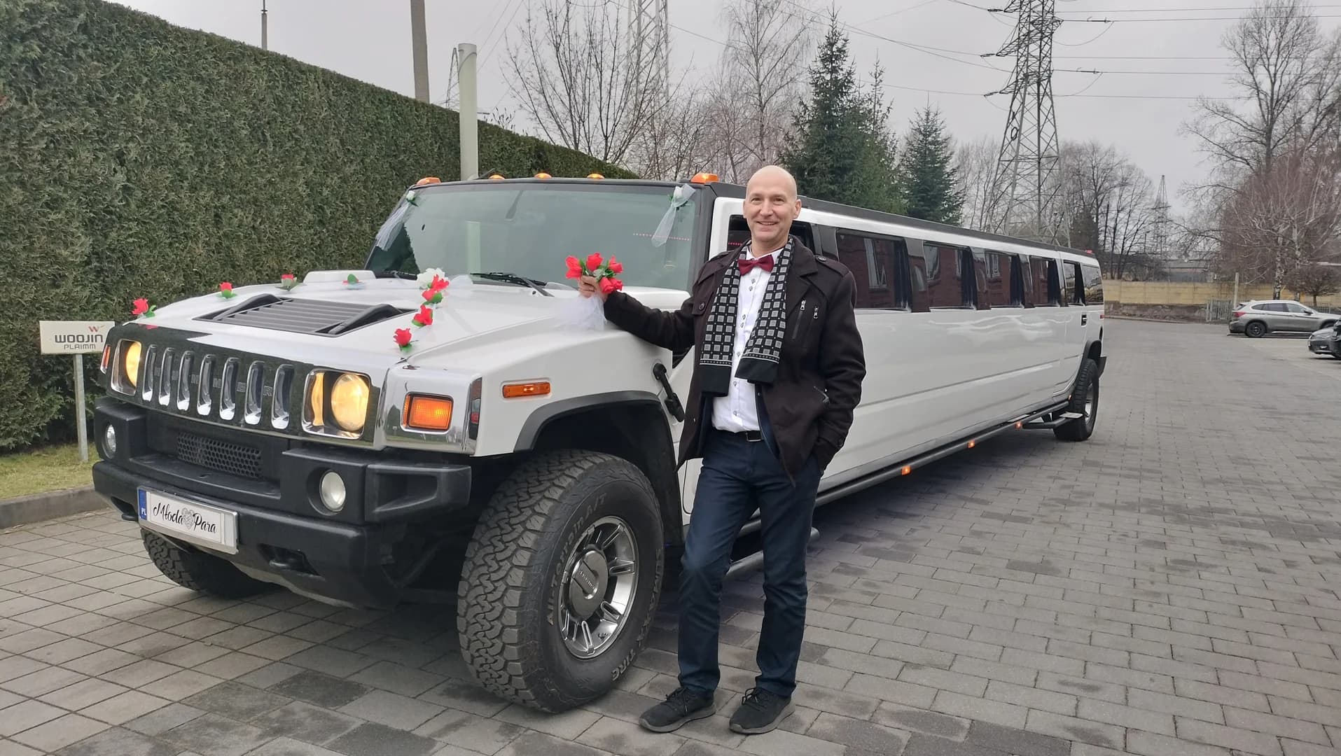 Mężczyzna w eleganckim stroju pozujący przy udekorowanej kwiatami białej limuzynie Hummer na parkingu.