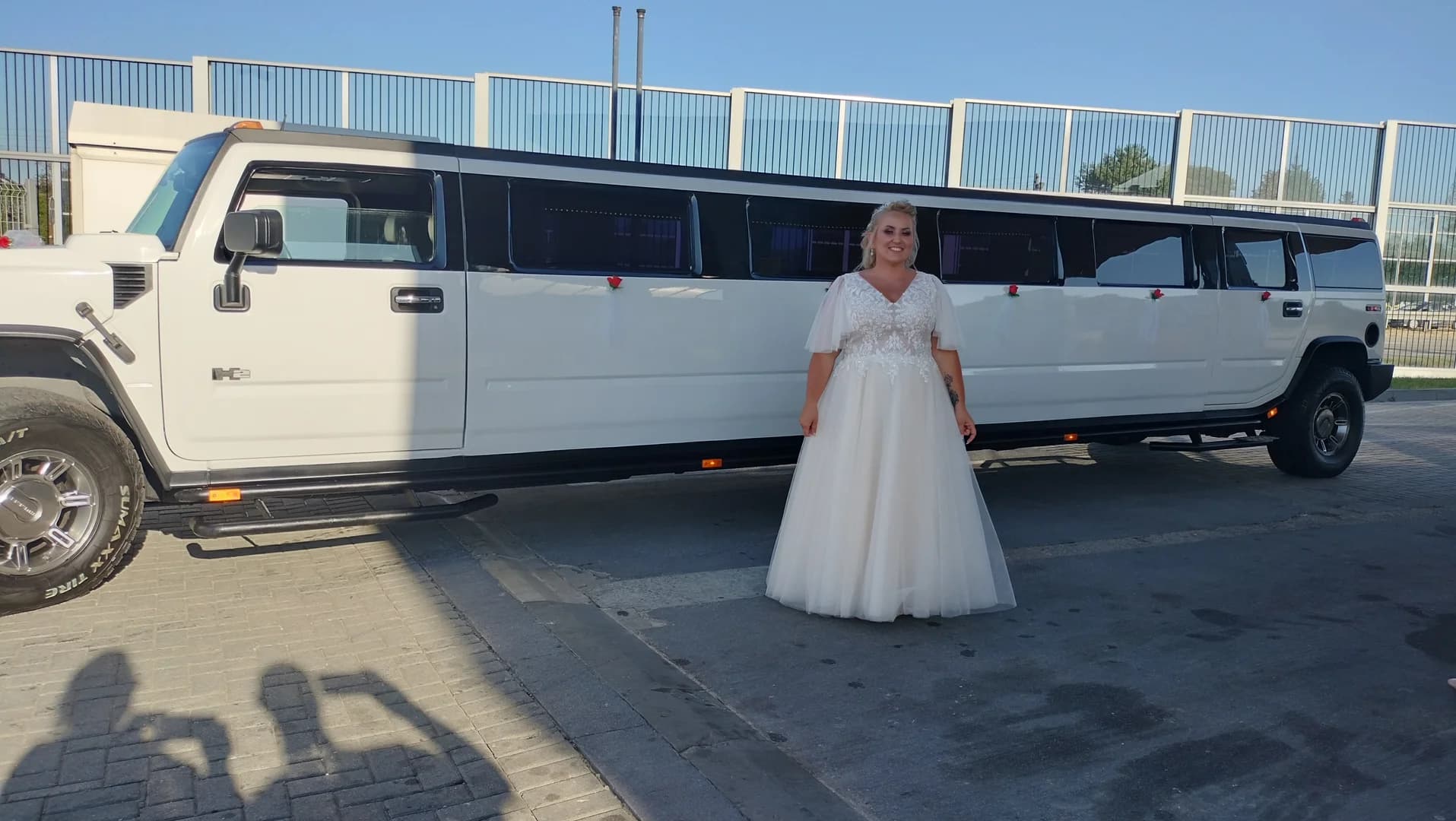 Panna młoda w białej sukni pozuje przed długą, białą limuzyną typu Hummer.
