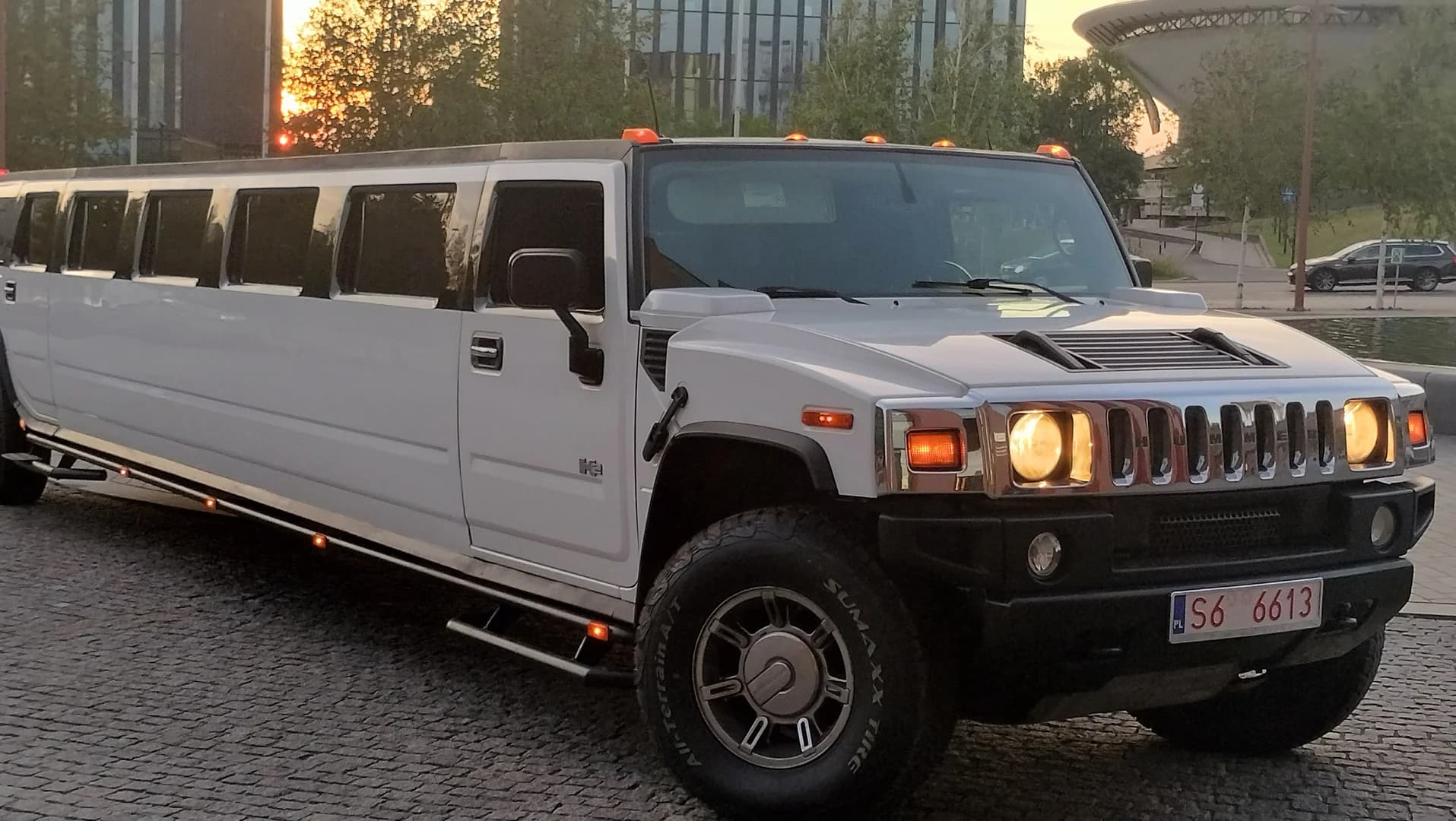 Biała limuzyna marki Hummer z włączonymi światłami zaparkowana na wybrukowanym placu o zachodzie słońca.