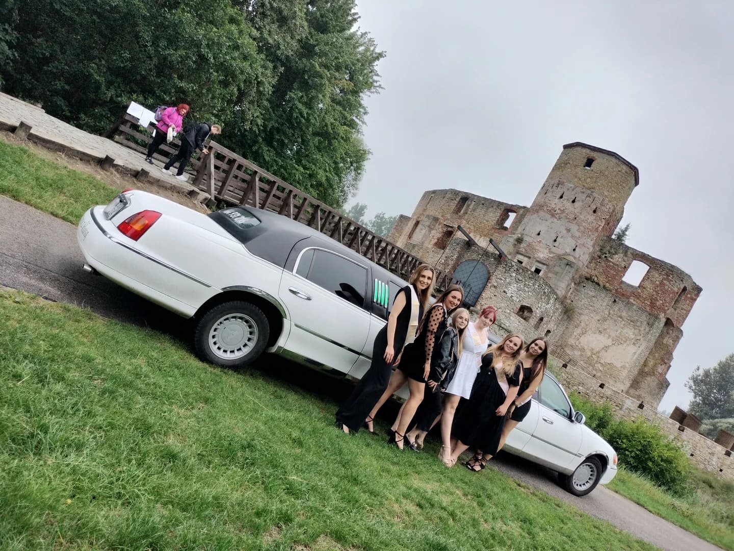 Grupa elegancko ubranych kobiet pozuje przed białą limuzyną w pobliżu zabytkowych ruin zamku.