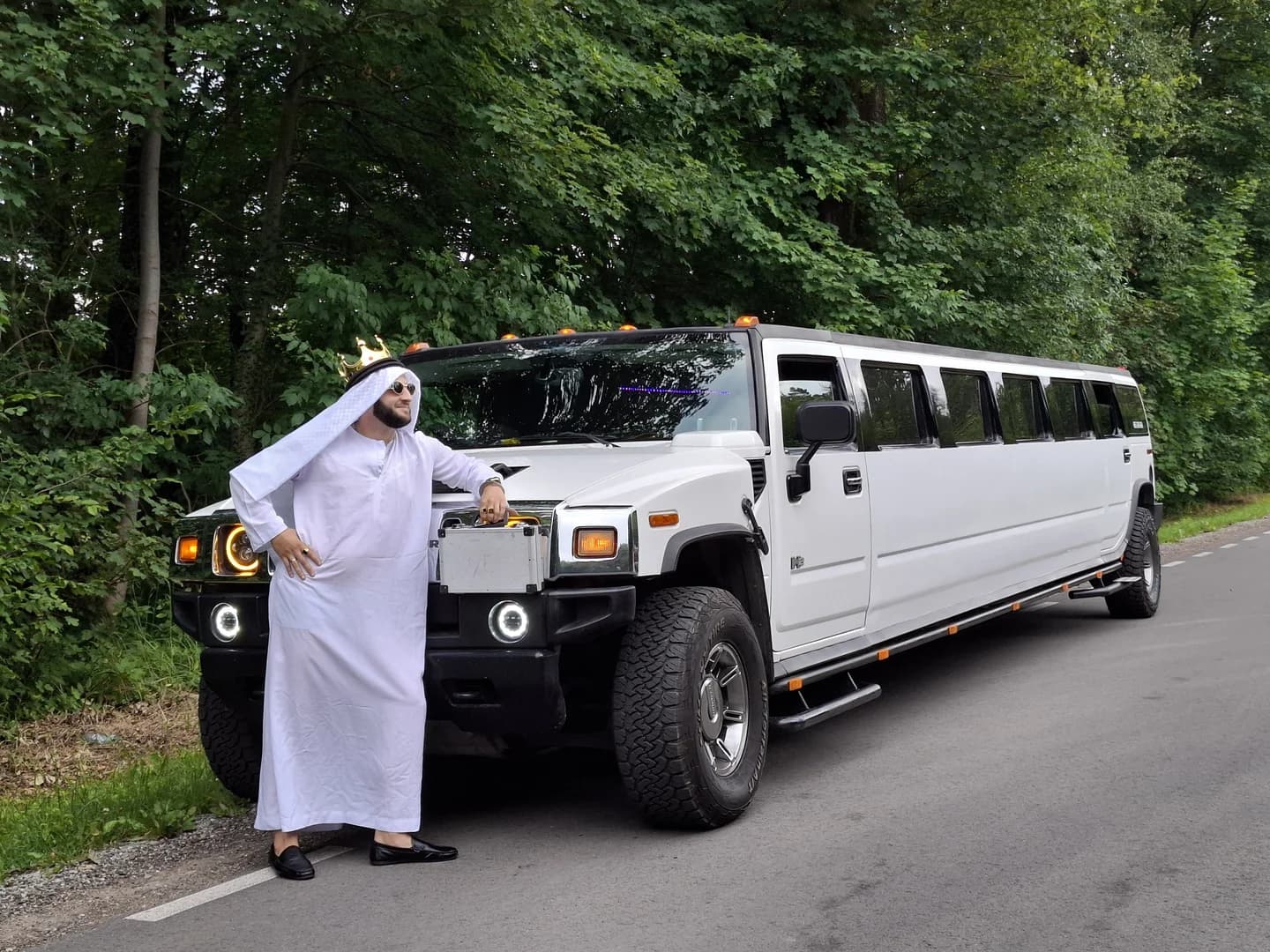 Mężczyzna w białym tradycyjnym stroju arabskim oraz koronie na głowie pozujący przy białej limuzynie Hummer zaparkowanej na skraju drogi przy lesie.