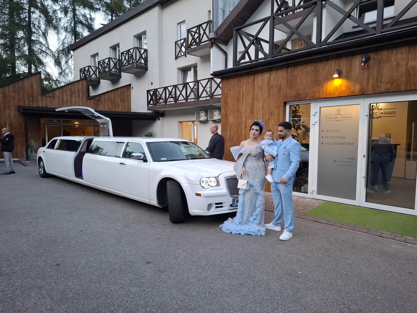 Elegancka rodzina w błękitnych strojach stoi przy długiej, białej limuzynie przed gościńcem.