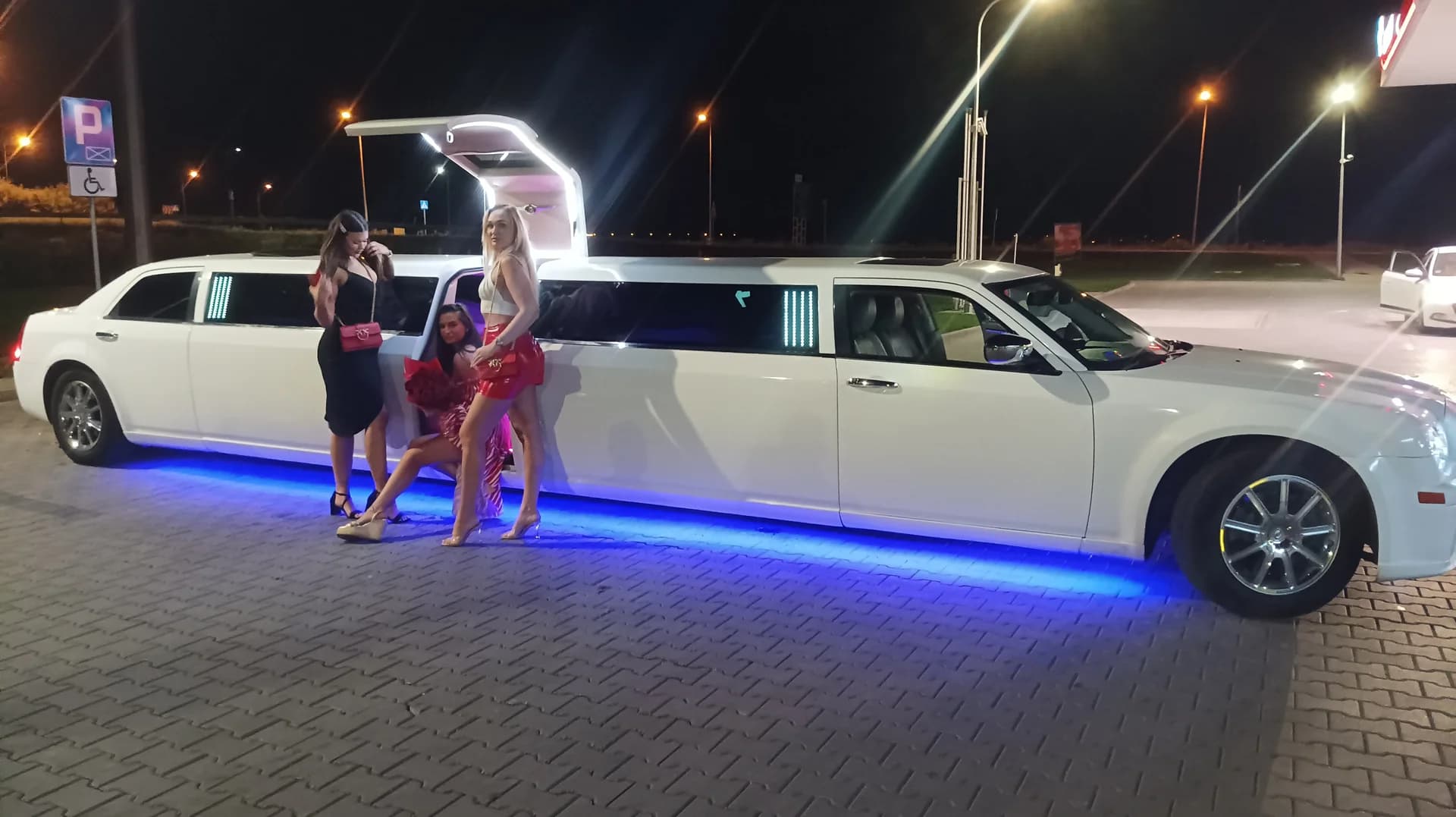 Trzy kobiety pozują nocą przy białej, podświetlanej na niebiesko limuzynie z otwartymi drzwiami.