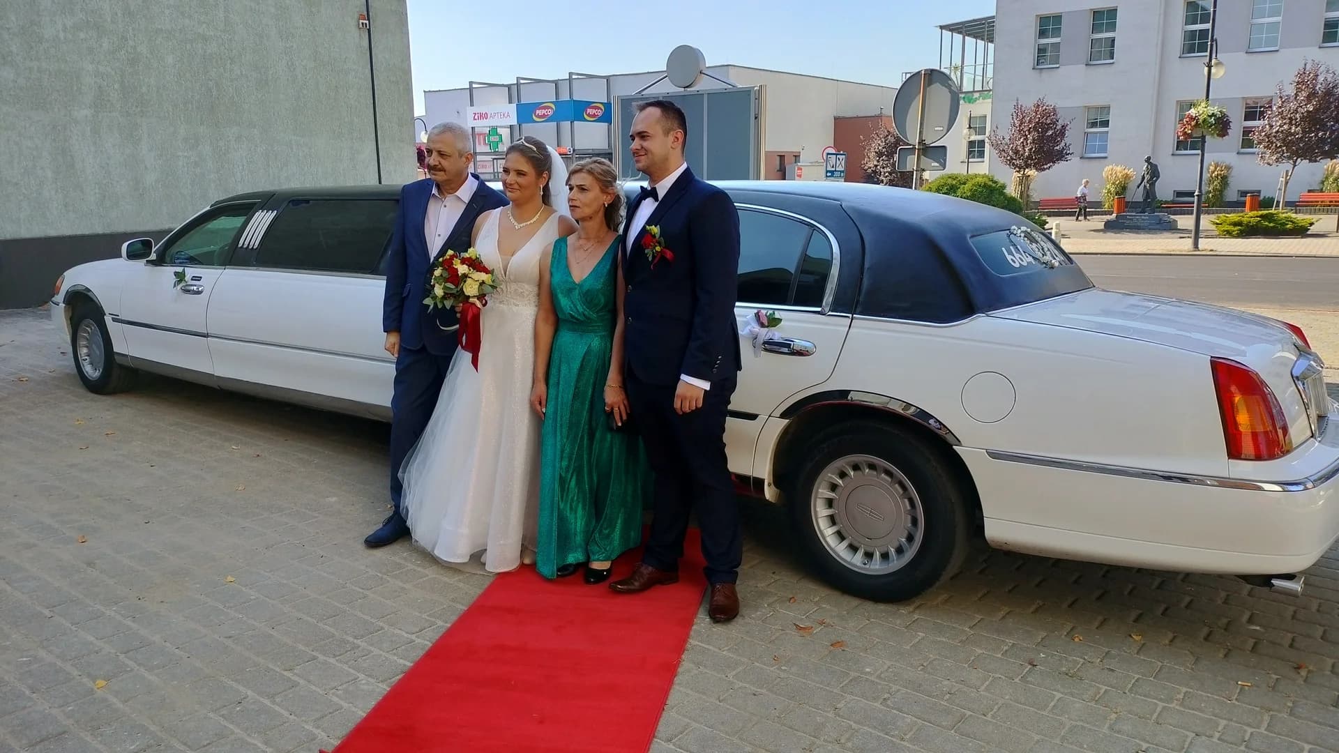 Czworo ludzi pozujących na czerwonym dywanie przy białej limuzynie ślubnej w słoneczny dzień.