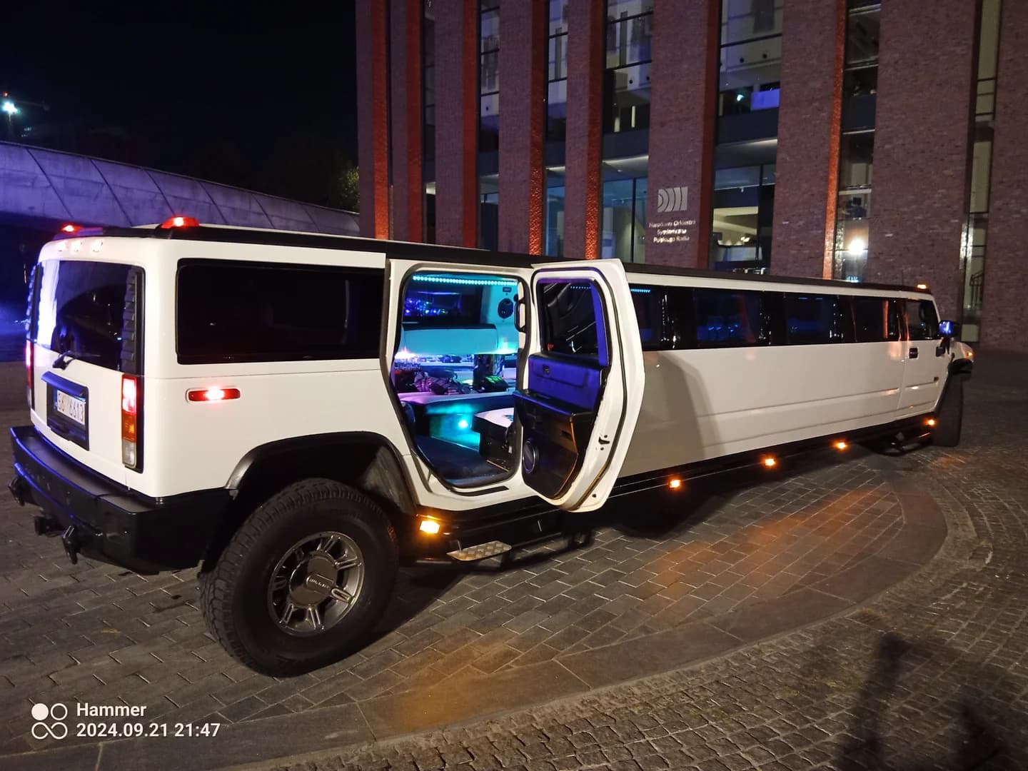 Biała limuzyna typu Hummer z otwartymi drzwiami i widocznym podświetlanym wnętrzem, zaparkowana wieczorem na brukowanym placu przed nowoczesnym budynkiem.
