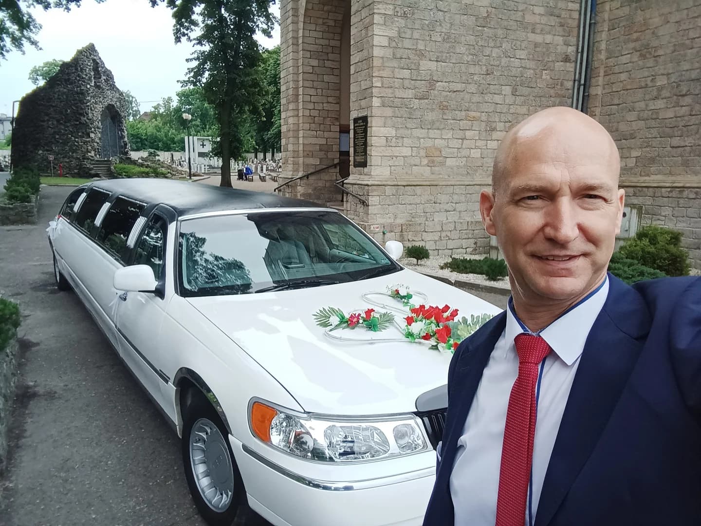 Mężczyzna w garniturze robi selfie na tle białej, ślubnej limuzyny przed kościołem.
