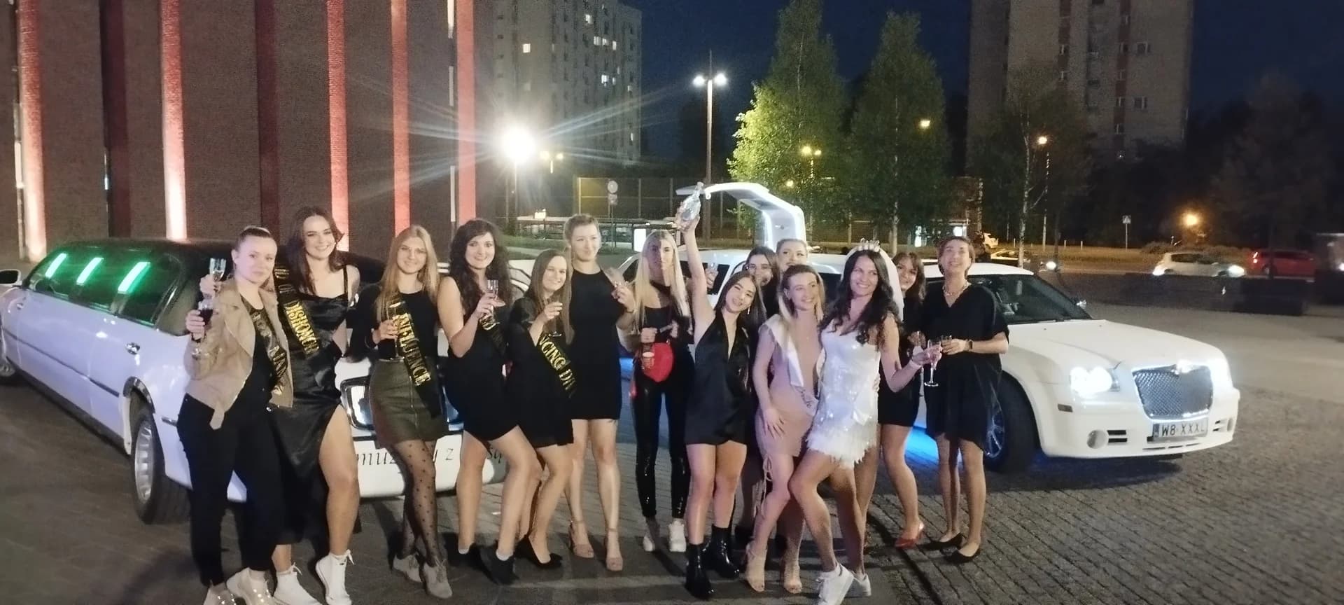 Duża grupa kobiet świętuje w nocy na tle dwóch białych limuzyn.