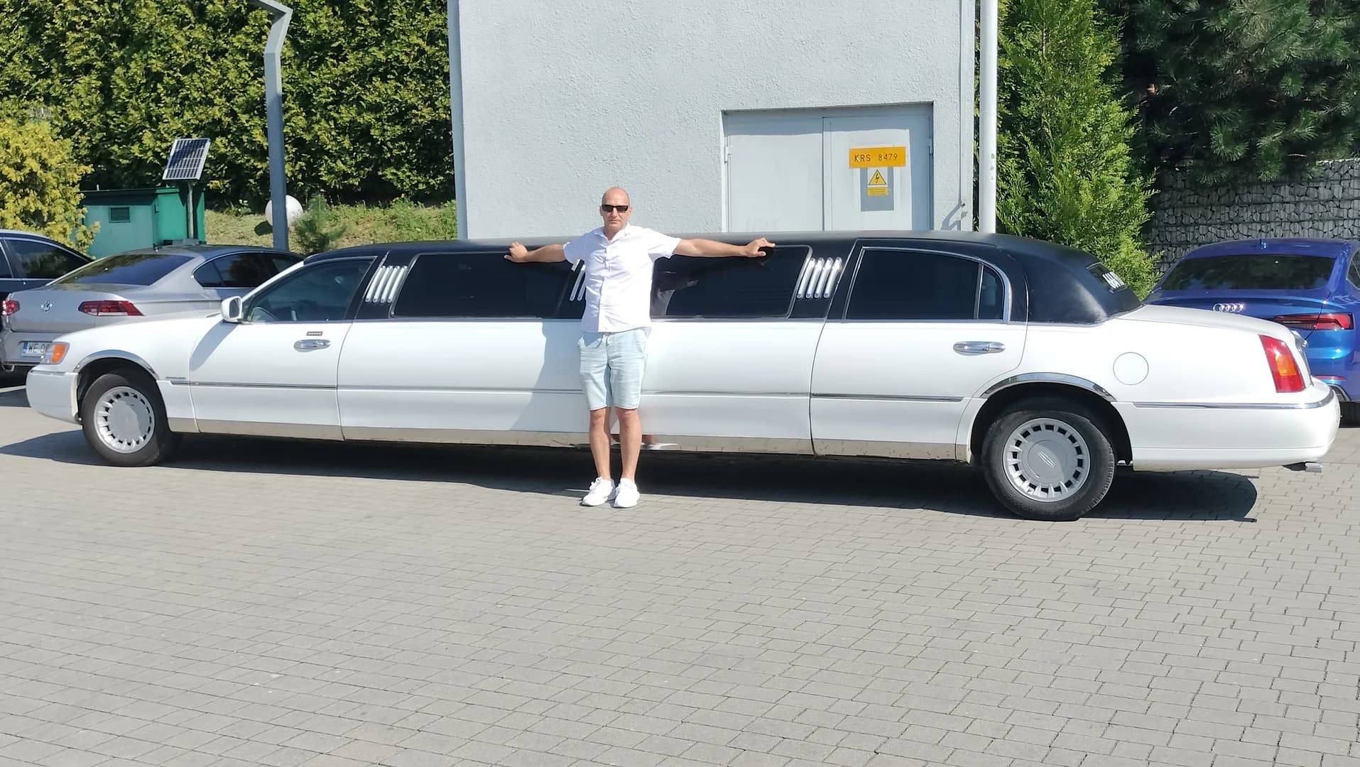Mężczyzna w jasnym stroju opiera się o bardzo długą, białą limuzynę na parkingu.