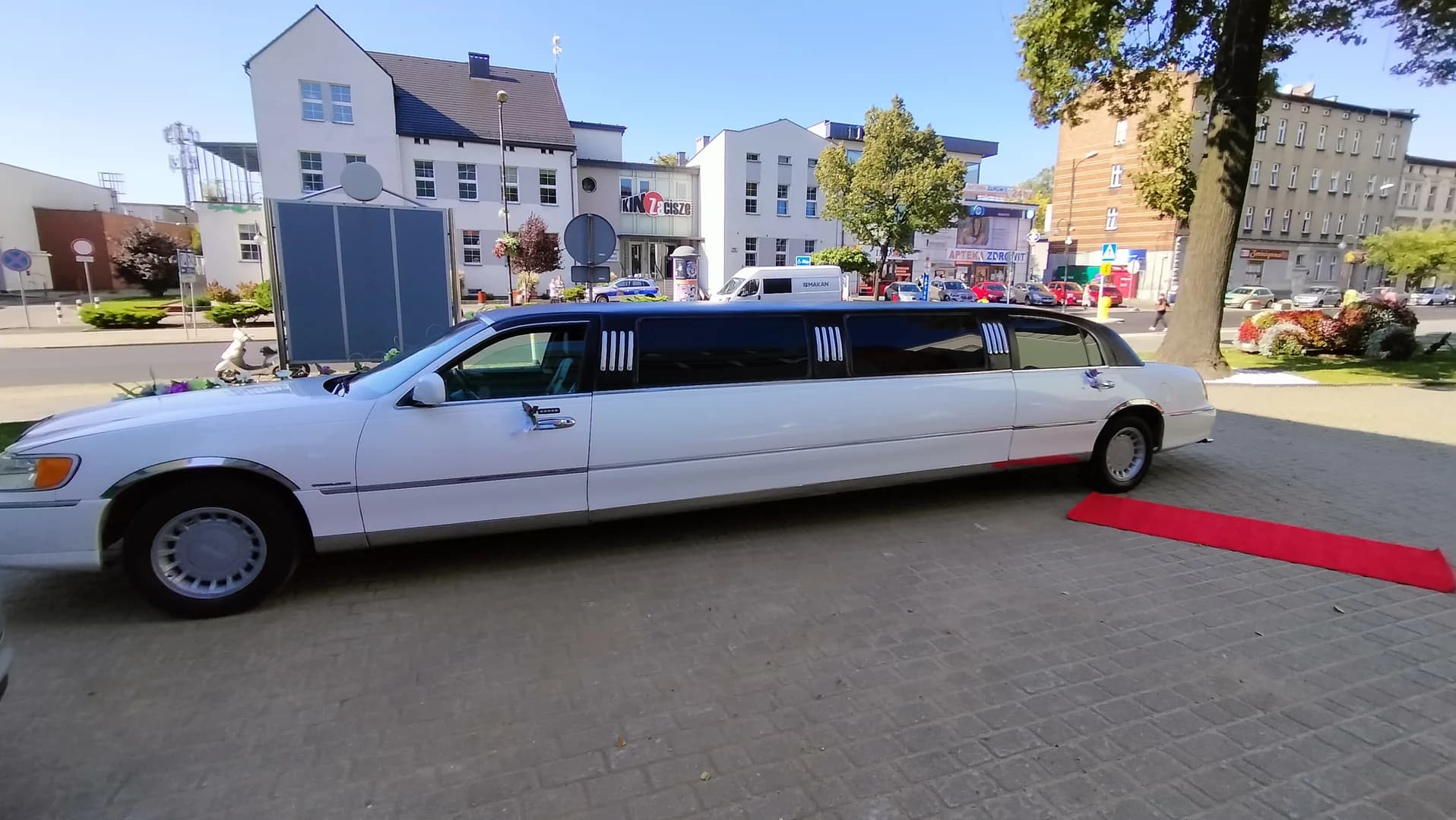 Biała limuzyna zaparkowana na brukowanym placu obok rozwiniętego czerwonego dywanu, w tle widoczna miejska zabudowa.