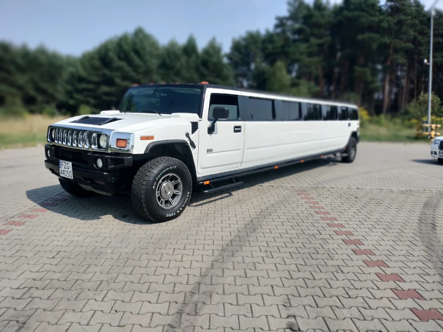 Biała limuzyna typu Hummer zaparkowana na wybrukowanym placu w słoneczny dzień.